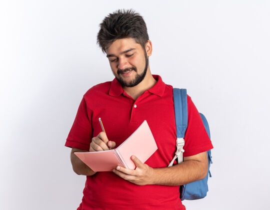 小伙子年轻的留着胡子的学生 穿着红色马球衫 背着书包 站在笔记本上写着自信的微笑笔记本微笑红色