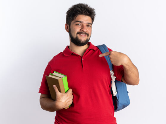 手指快乐的年轻留着胡子的学生 穿着红色马球衫 背着背包 拿着笔记本 用食指指着笔记本 开心地站着红色快乐欢呼