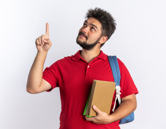 年轻年轻的留着胡子的学生 穿着红色马球衫 背着背包 拿着笔记本 微笑着抬起头 用食指指着站着的东西红色拿着手指