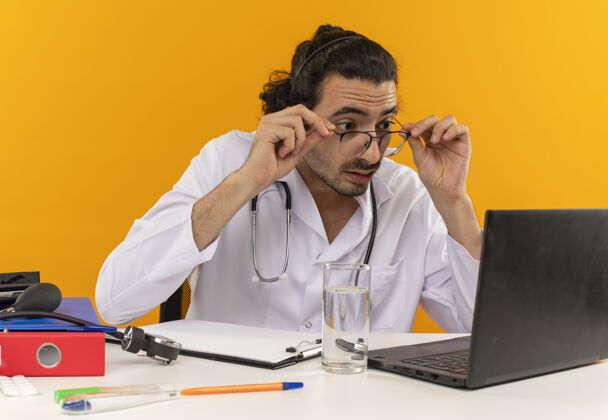 坐着惊讶的年轻男医生戴着医用眼镜 穿着医用长袍 听诊器坐在办公桌旁眼镜工作长袍