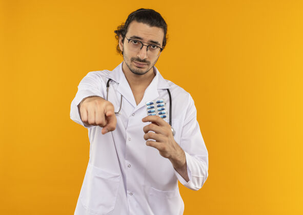 医疗悲伤的年轻医生戴着医用眼镜穿着医用长袍手持听诊器拿着药片给你看手势持有手势药丸