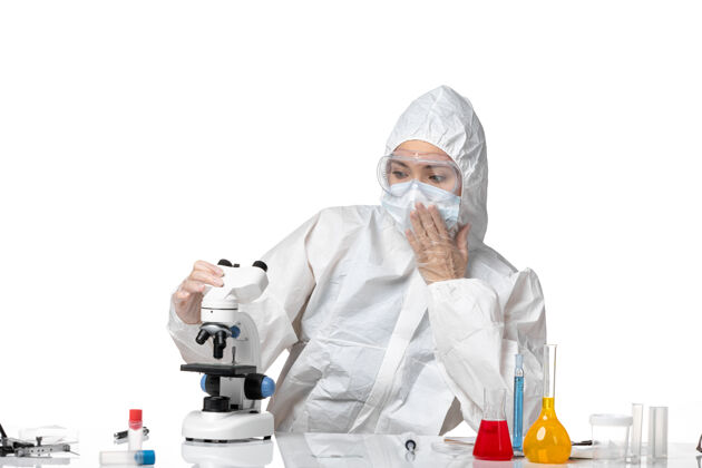 药品正面图年轻女医生穿着白色防护服 戴着口罩 由于使用显微镜在白色办公桌上大流行 飞溅病毒健康使用外套医生