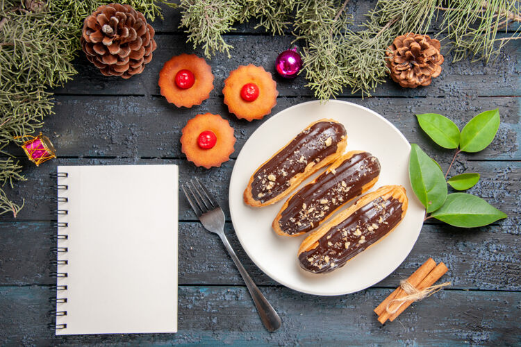 茶俯瞰图：白色椭圆形平板上的巧克力蛋糕 冷杉树枝和圆锥体 圣诞玩具 叉子 肉桂 一杯茶 黑木桌上的笔记本树枝玩具圣诞节