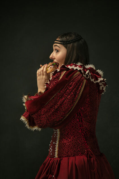 好激情四射身着红色复古服装的中世纪年轻女子在深色背景下吃汉堡包的肖像女模特是公爵夫人 皇室成员时代 现代 时尚 美丽的对比概念历史人服装