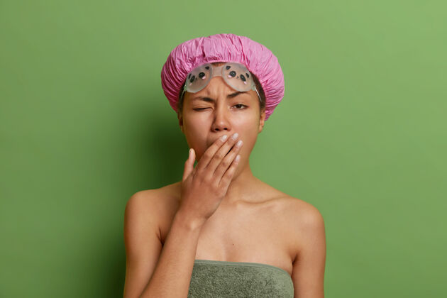毛巾困倦的美国黑人妇女用手捂住嘴想要睡觉裹着浴巾戴着防水帽游泳镜隔着鲜艳的绿色墙壁健康早晨美丽