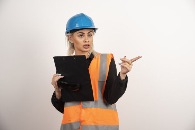 人类年轻的女建筑工人用剪贴板指向某处高质量的照片头盔建筑女士