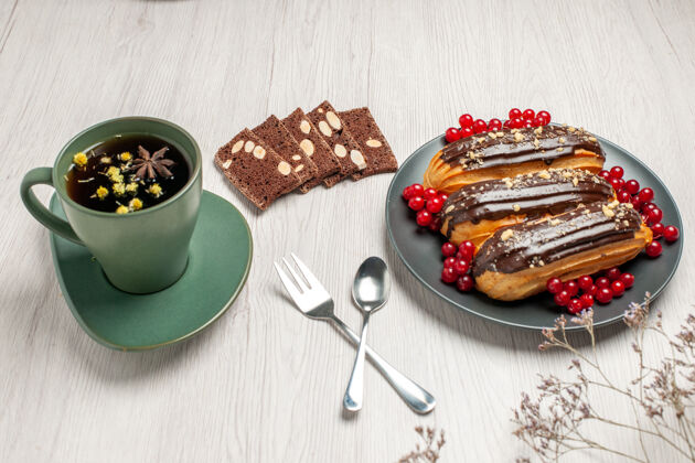 叉子底视图巧克力eclairs和黑醋栗在灰色板花生饼干一杯茶勺子和叉子在对角线向量在白色的木桌上晚餐胡椒杯子