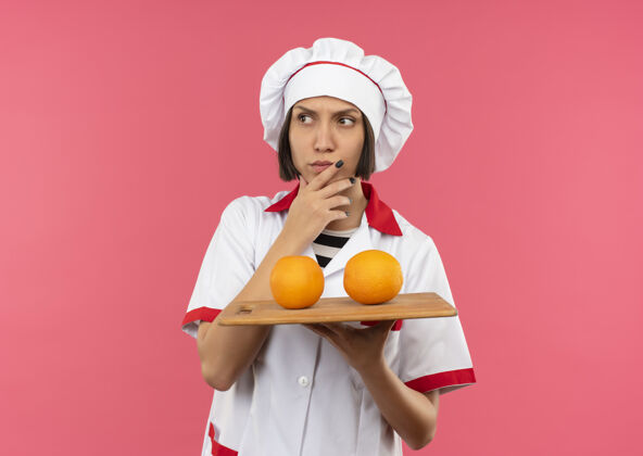 女性体贴的年轻女厨师穿着厨师制服 拿着砧板 砧板上放着桔子 抚摸着下巴 看着粉红色墙上孤立的一面橙色拿着下巴