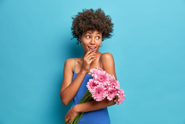 花可爱女人的肖像 卷发 穿着节日服装 手持一束非洲菊花 在第一次约会时 她高兴地站在一边 隔着蓝色的墙壁美丽女性服装