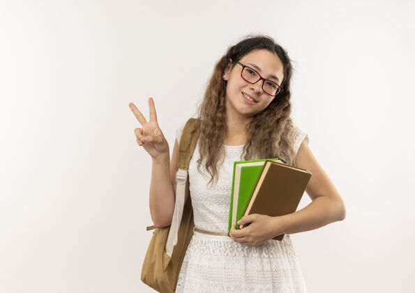 脸微笑着的年轻漂亮的女学生戴着眼镜背着书包在白色的墙上做着和平的标志和平感情人