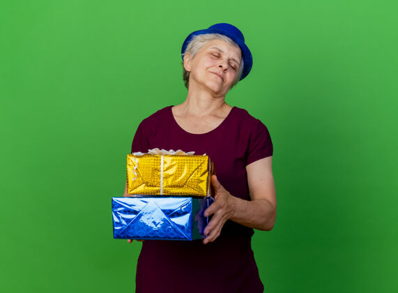 女士高兴的老妇人戴着聚会帽闭着眼睛拿着礼品盒上的绿色请关闭礼物帽子