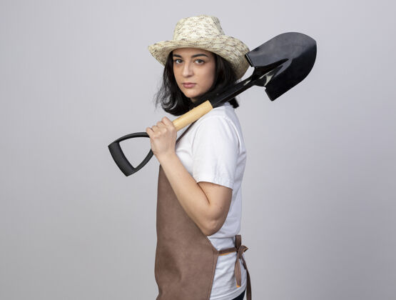 举行自信的年轻黑发女园丁穿着制服 戴着园艺帽 站在一旁 手里拿着黑桃制服信心黑桃