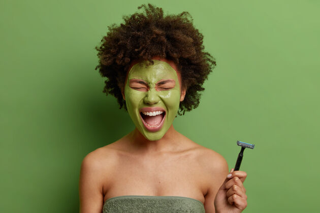 卫生情绪化的卷发年轻女子裹着柔软的浴巾拿着剃须刀去脱毛敷上美容面膜治疗皮肤保持张大嘴巴隔离绿色墙壁粘土过程面具
