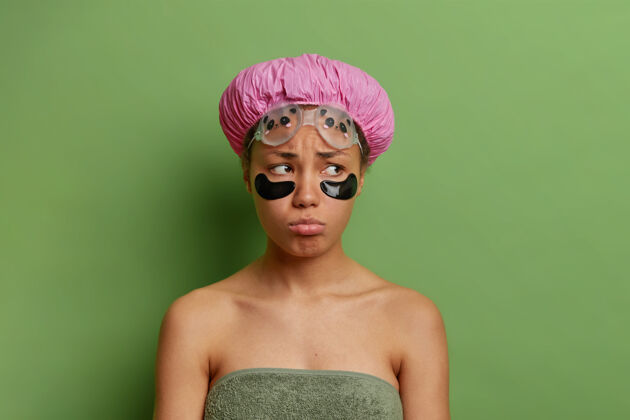 肩膀愁眉苦脸的女人洗完澡 裹着毛巾 戴着隔离在绿墙上的浴帽 进行美容手术水疗胶原蛋白人类