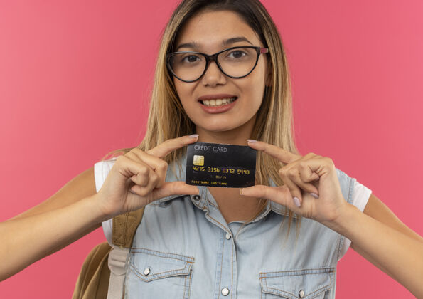 包面带微笑的年轻漂亮的女学生戴着眼镜 背着一个袋子 手里拿着信用卡 就在前面的粉红色墙上微笑人感情