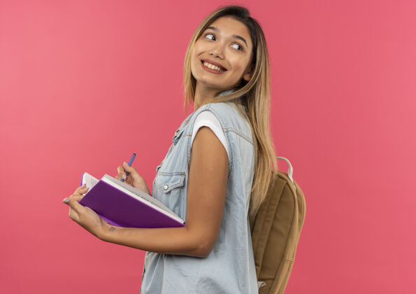 粉色面带微笑的年轻漂亮的学生女孩 背着书包站在侧视图中 手里拿着打开的书和笔 看着身后孤立的粉色墙壁开脸感觉