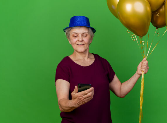 拜托戴着派对帽的老太太高兴地拿着氦气球 看着绿色的手机女人老人派对