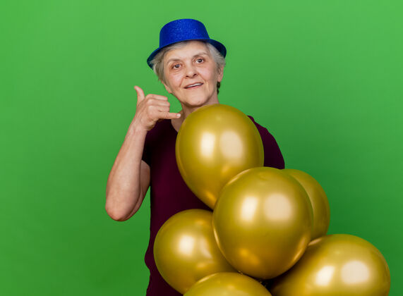 气球高兴的老妇人戴着党的帽子站在氦气球手势叫我绿色的标志呼叫老人帽子