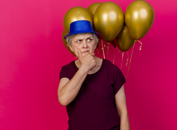 困惑困惑的老妇人戴着派对帽 在氦气球前托着下巴看着粉色的一面气球派对前面