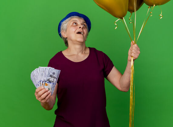 气球戴着派对帽的兴奋的老妇人手里拿着钱 看着绿色的氦气球兴奋帽子女人