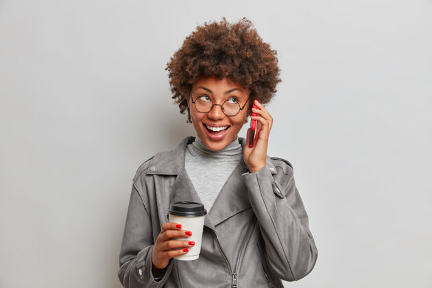 高兴工作室拍摄的积极可爱的年轻黑人美国妇女愉快的交谈保持智能手机近耳饮料外卖咖啡隔离灰色的墙壁满意姿势请