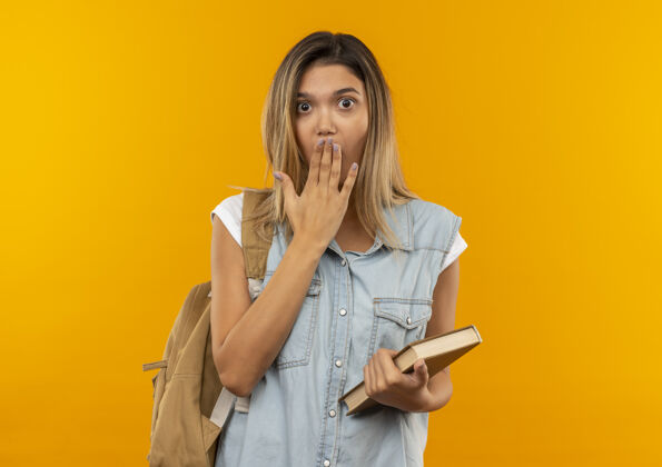 表情惊讶的年轻漂亮的女学生穿着背袋拿着书 把手放在嘴巴上孤立在橙色的墙上抱背橘子