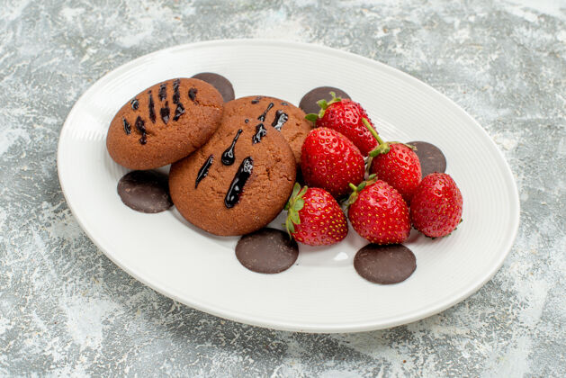盘子特写俯视图巧克力饼干草莓和圆形巧克力在白色椭圆形盘子上的灰白色地面饮食椭圆形磨碎