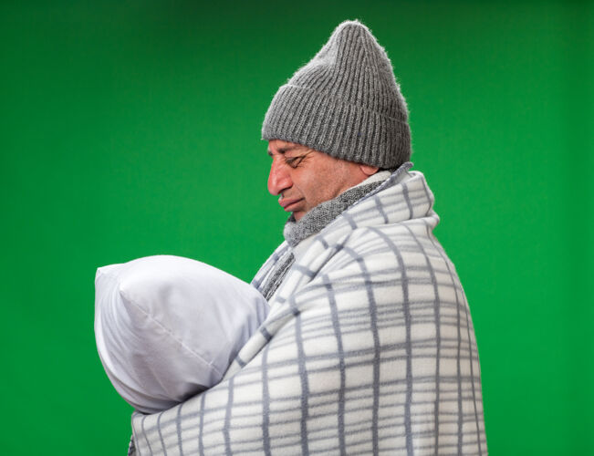 格子悲伤的成年白种人 脖子上围着围巾 戴着格子布裹着的冬日帽 抱着一个枕头 看着隔离在绿色墙壁上的复制空间帽子枕头成人