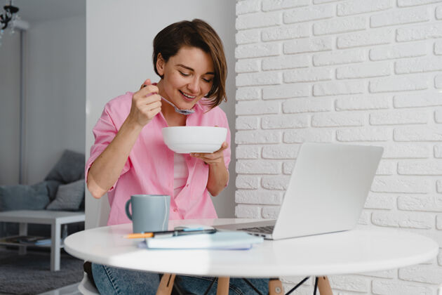 Muesli穿着粉色衬衫的微笑女人在家里吃早餐在桌边用笔记本电脑上网吃饭笔记本电脑坐着