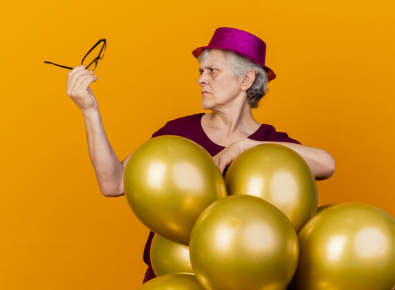 表情无知的老妇人戴着聚会帽站在那里 手里拿着氦气球 看着隔离在橙色墙上的眼镜人女人人