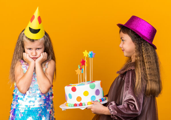 悲伤高兴的小白种女孩戴着紫色派对帽拿着生日蛋糕 看着悲伤的小金发女孩戴着派对帽孤立地贴在橙色的墙上 还有复制空间金发孤立请