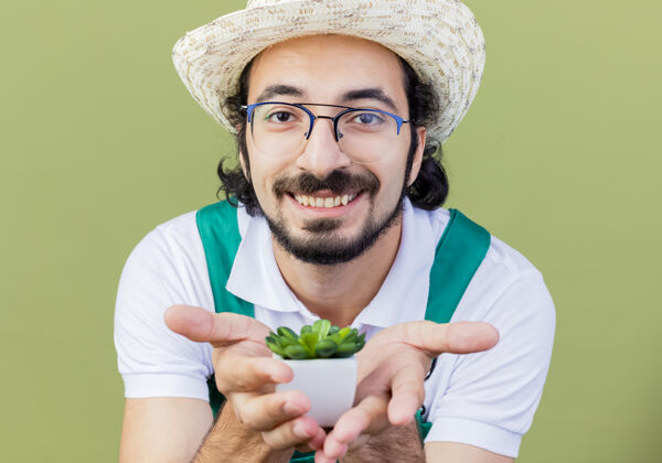 表演年轻的留着胡须的园丁 穿着连身衣 戴着帽子 展示着盆栽植物 站在浅绿色的墙上 面带微笑地看着前方市民花园人