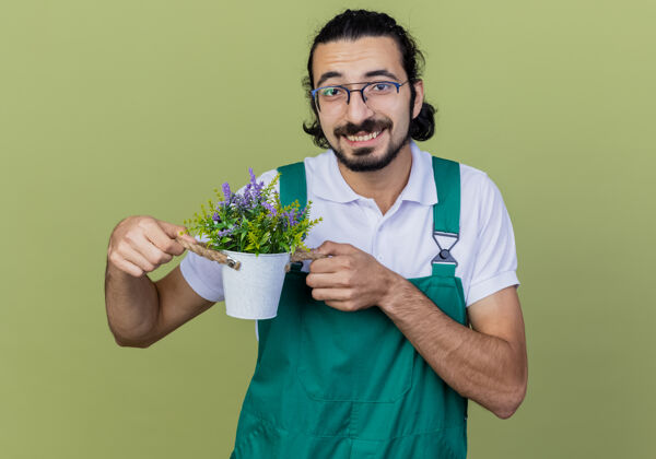 光年轻的留着胡须的园丁穿着连身衣 展示着盆栽植物 微笑着看着站在浅绿色墙上的前面年轻人胡须衣服