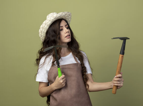 人自信的年轻女园丁穿着制服 戴着园艺帽 手持锄头耙 看着橄榄绿墙上孤立的耙子穿帽子脸