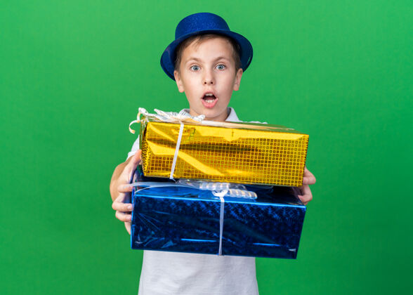 拿着惊讶的年轻斯拉夫男孩 戴着蓝色派对帽 拿着礼品盒 隔离在绿墙上 留着复印空间斯拉夫人盒子惊喜