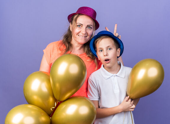 惊喜惊讶的年轻斯拉夫男孩 戴着蓝色的派对帽 手里拿着氦气球 母亲戴着紫色的派对帽 隔离在紫色的墙上 留着复制空间拿着男孩穿
