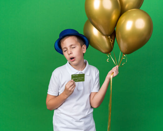 男孩恼怒的年轻斯拉夫男孩 戴着蓝色派对帽 手里拿着氦气球 信用卡被隔离在绿墙上 留着复印空间年轻帽子恼怒