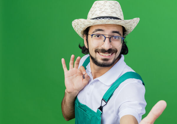 人年轻的留着胡须的园丁 穿着连体衣 戴着帽子 面带微笑地看着前面 绿色的墙上挂着“ok”的牌子男人表演站立