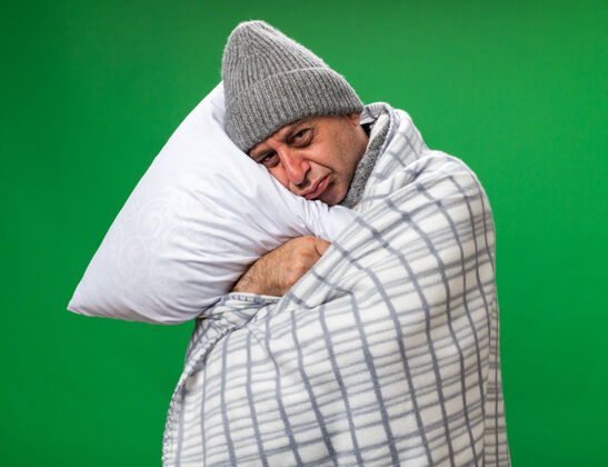 头悲伤的成年病高加索男子脖子上围着围巾 戴着格子布包裹的冬季帽子 抱着头枕在枕头上 隔离在绿色墙壁上 留着复制空间围着裹着冬天