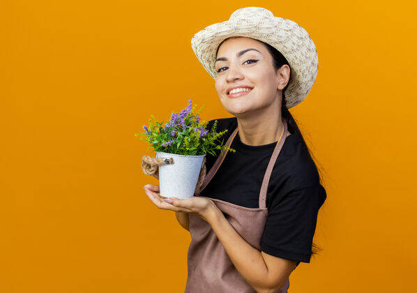 漂亮年轻漂亮的女园丁围着围裙 戴着帽子 手里拿着盆栽植物 站在橘色的墙上微笑着看着前方帽子花园市民