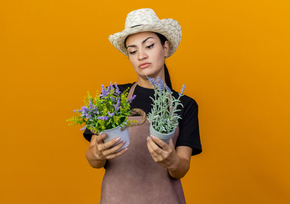 站年轻漂亮的女园丁 围着围裙 戴着帽子 手里拿着盆栽植物 站在橘红色的墙上困惑地看着它们壶衣服帽子