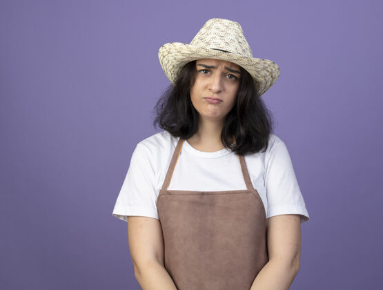 表情悲伤的年轻黑发女园丁穿着制服 戴着园艺帽 看着隔离在紫色墙上的前面帽子人人