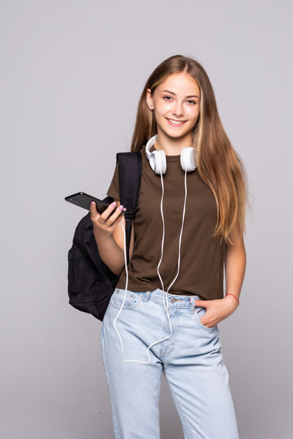 成人带着智能手机的年轻女子隔着白墙上的背包听音乐背包人耳机
