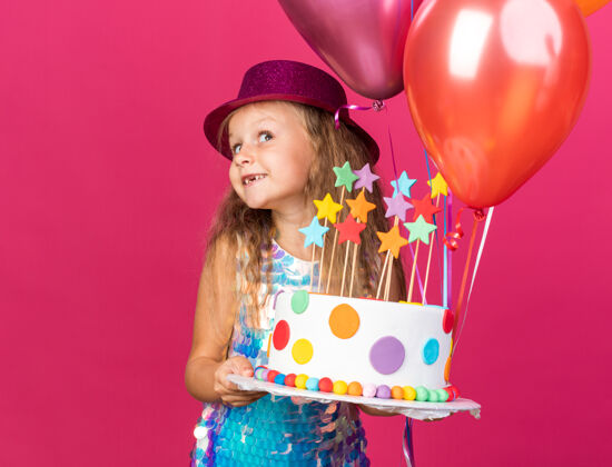 氦可爱的金发小女孩 戴着紫色的派对帽 手里拿着氦气球和生日蛋糕 隔离在粉红色的墙上 还有复制空间拜托派对女孩