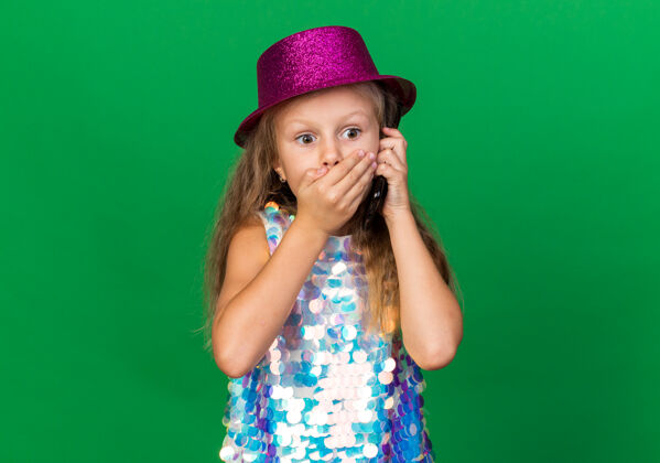 孤立震惊的金发小女孩 戴着紫色派对帽 手放在嘴上 讲着电话 隔离在绿色的墙上 留着复印空间派对金发帽子