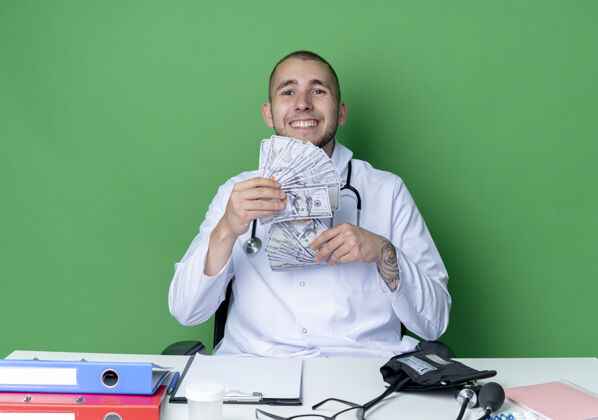 坐着面带微笑的年轻男医生穿着医用长袍和听诊器坐在办公桌旁 手里拿着工作工具 手里拿着隔离在绿墙上的钱衣服医生工具