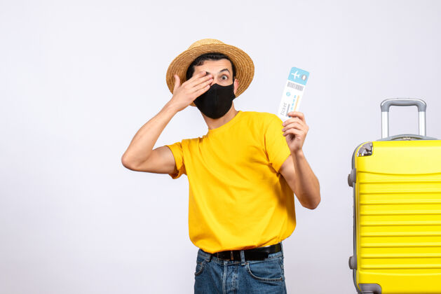 男人正面图身穿黄色t恤的年轻人站在黄色手提箱旁 手举旅行票遮住眼睛T恤人手