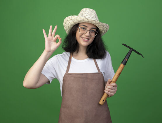 手年轻的深色皮肤的女园丁面带微笑 戴着眼镜和制服 戴着园艺帽 在绿色的墙上做着手势 拿着耙子穿着园艺帽子