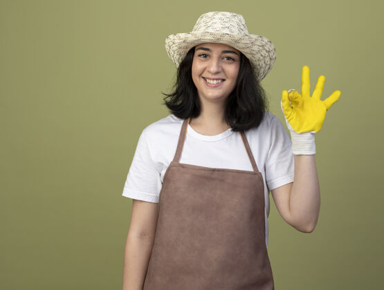 姿势微笑的年轻黑发女园丁穿着制服戴着园艺帽和手套手势ok手势隔离在橄榄绿的墙上人手套穿着