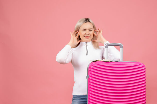 关闭正面图：站在粉色手提箱后面的年轻女士用手指合上耳朵成人女士站立